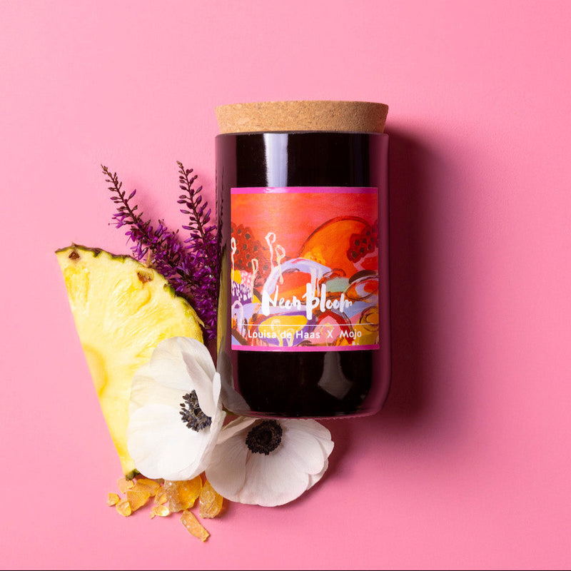 Candle with a colorful label on a pink background, surrounded by flowers and a pineapple slice.